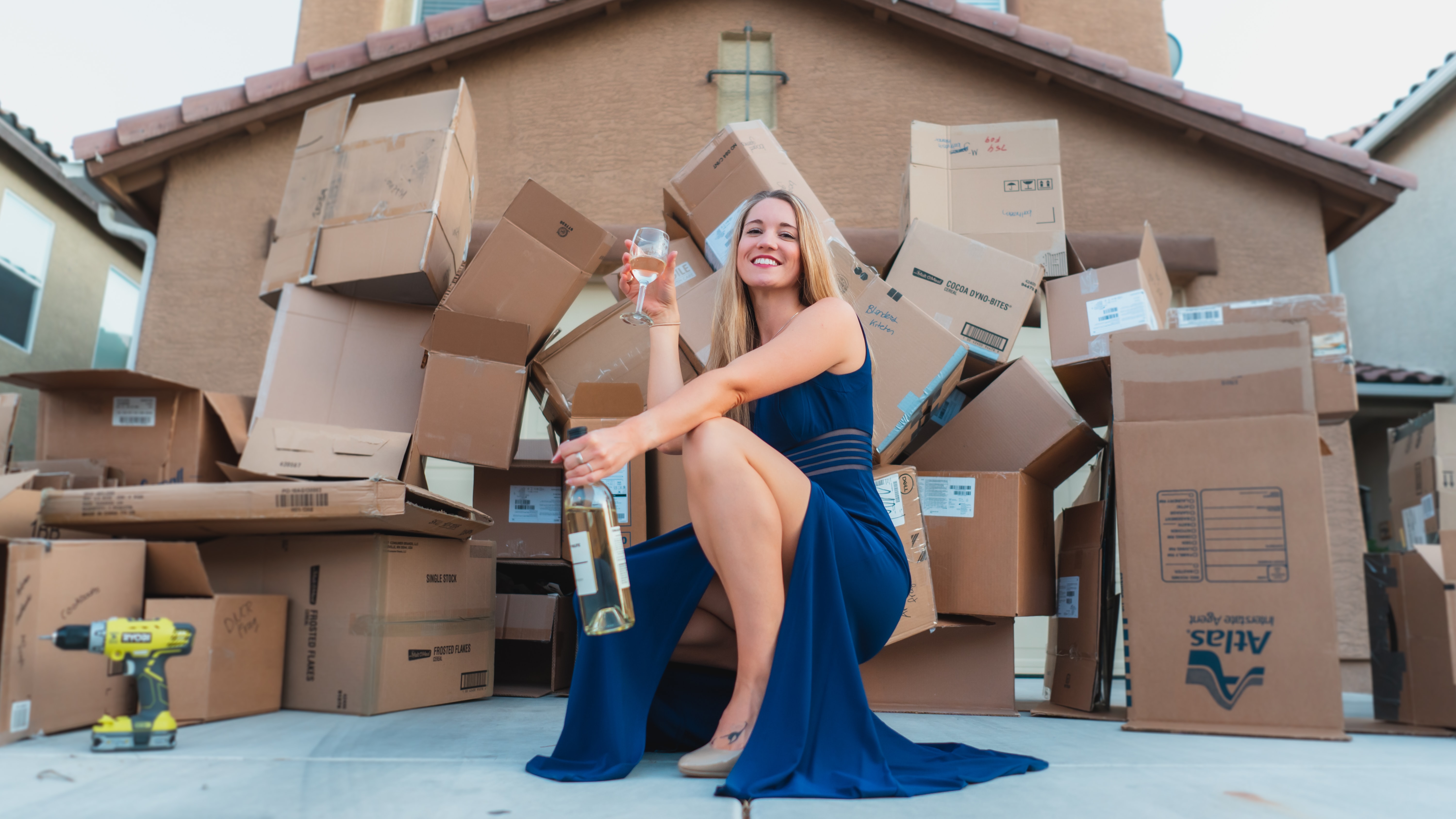 : A girl in an elegant blue dress standing in front of the boxes holding a bottle and a glass of wine